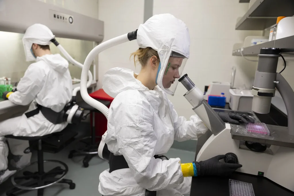 Two researchers in white bio-hazard suits observe samples in a lab. They are facing away from each other at different desks. One is looking through a microscope and the other looks at their desk.