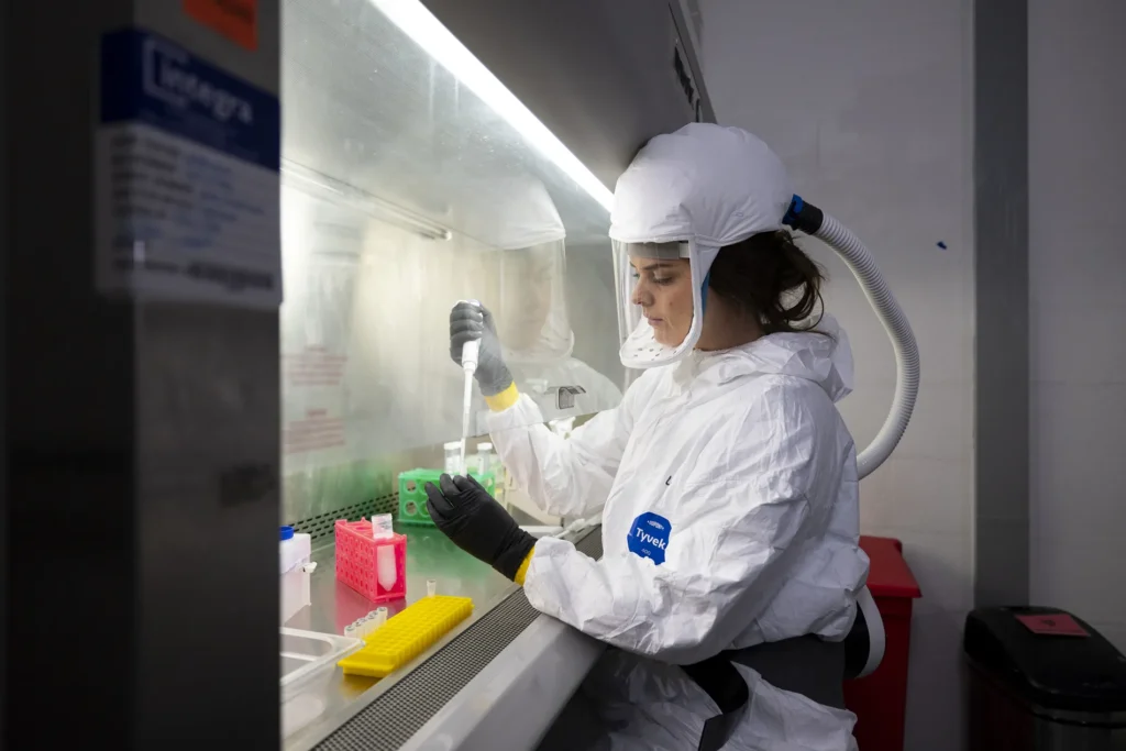 A researcher in a white bio-hazard suit and black gloves sits at a steel table with a bright florescent light overhead. In her right hand is a long tool that looks like an upside-down squeeze bottle. She uses it to put a sample in the test tube in her left hand.