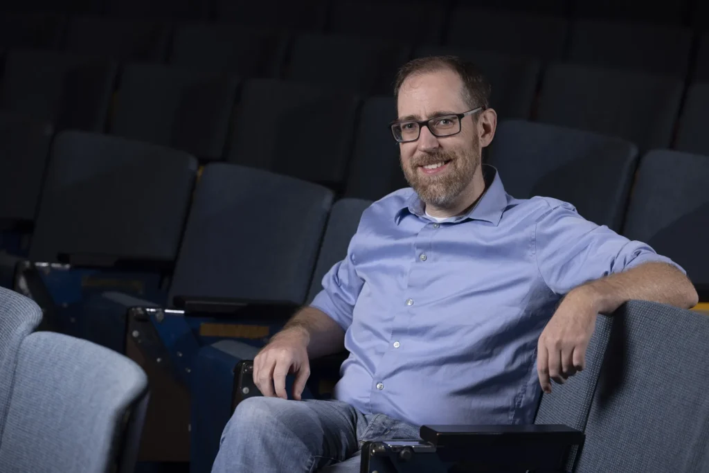 Profile of a man wearing a blue collared shirt and smiling at the camera. He is sitting in a theatre seat surrounded by empty seats.