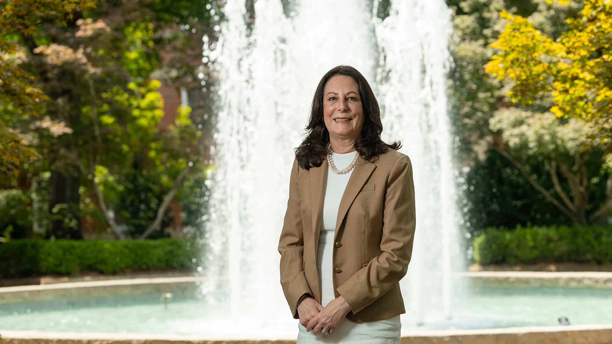 A woman in a tan jacket and white blouse poses in front of a large courtyard fountain.