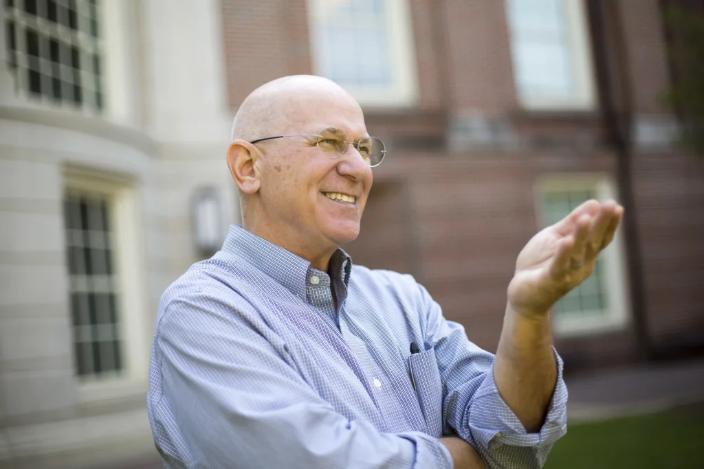 A closeup of a man in a blue-collared shirt smiling outside of an academic building.
