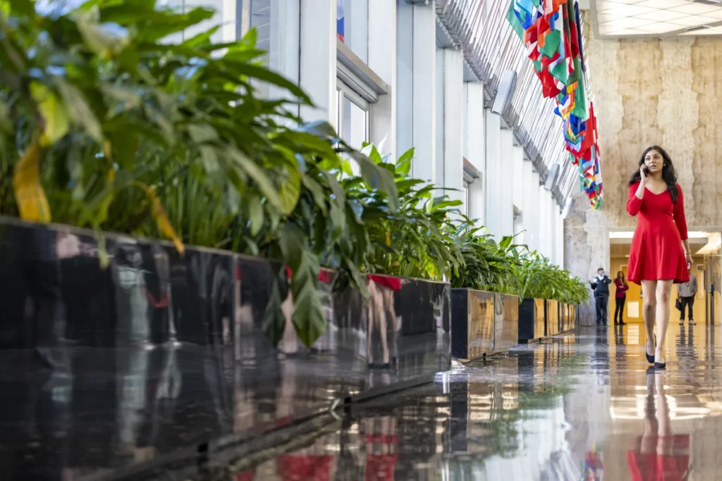 A young woman in a red dress walks down a brightly-lit hallway near a row of plants.