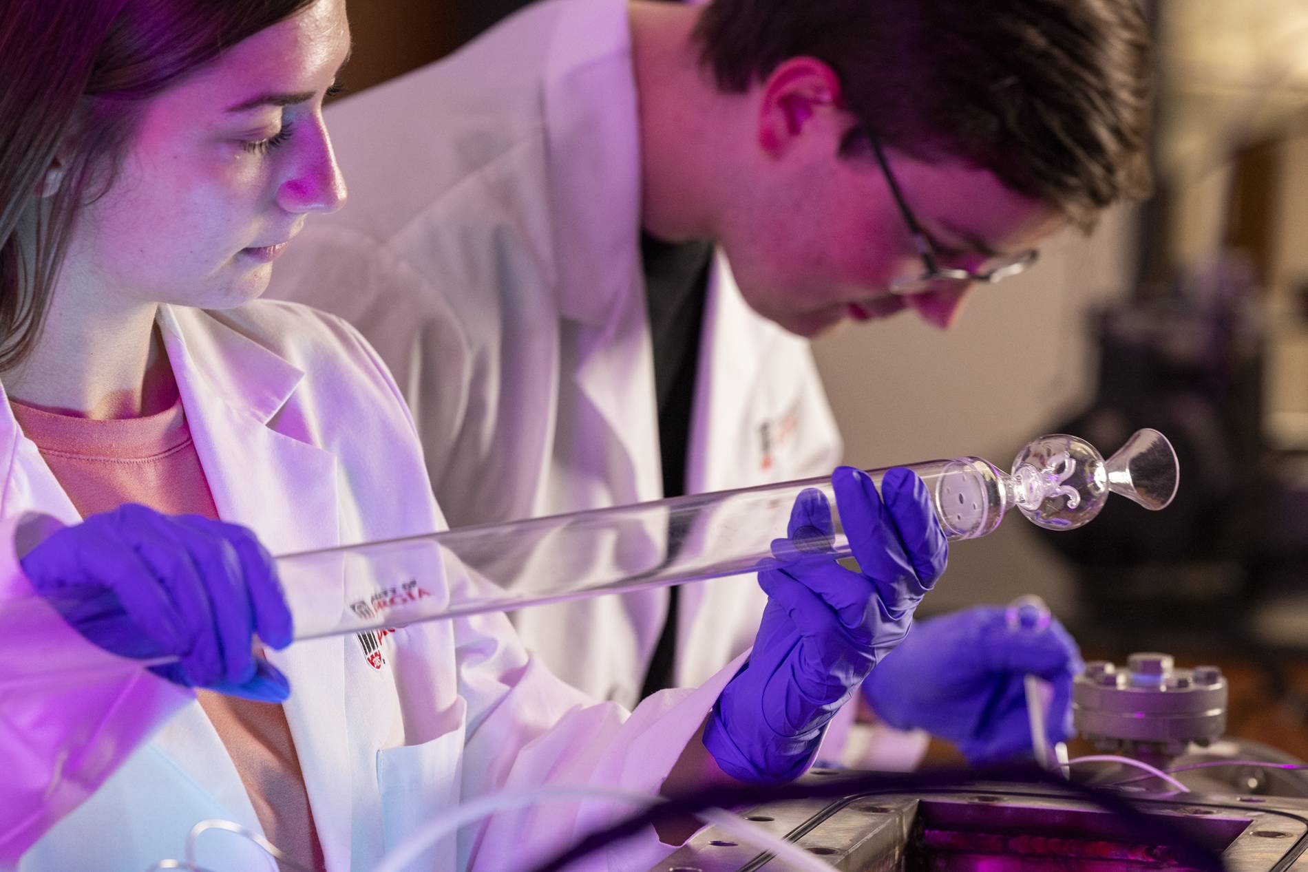 A closeup of two students in white lab coats looking at glass equipment.